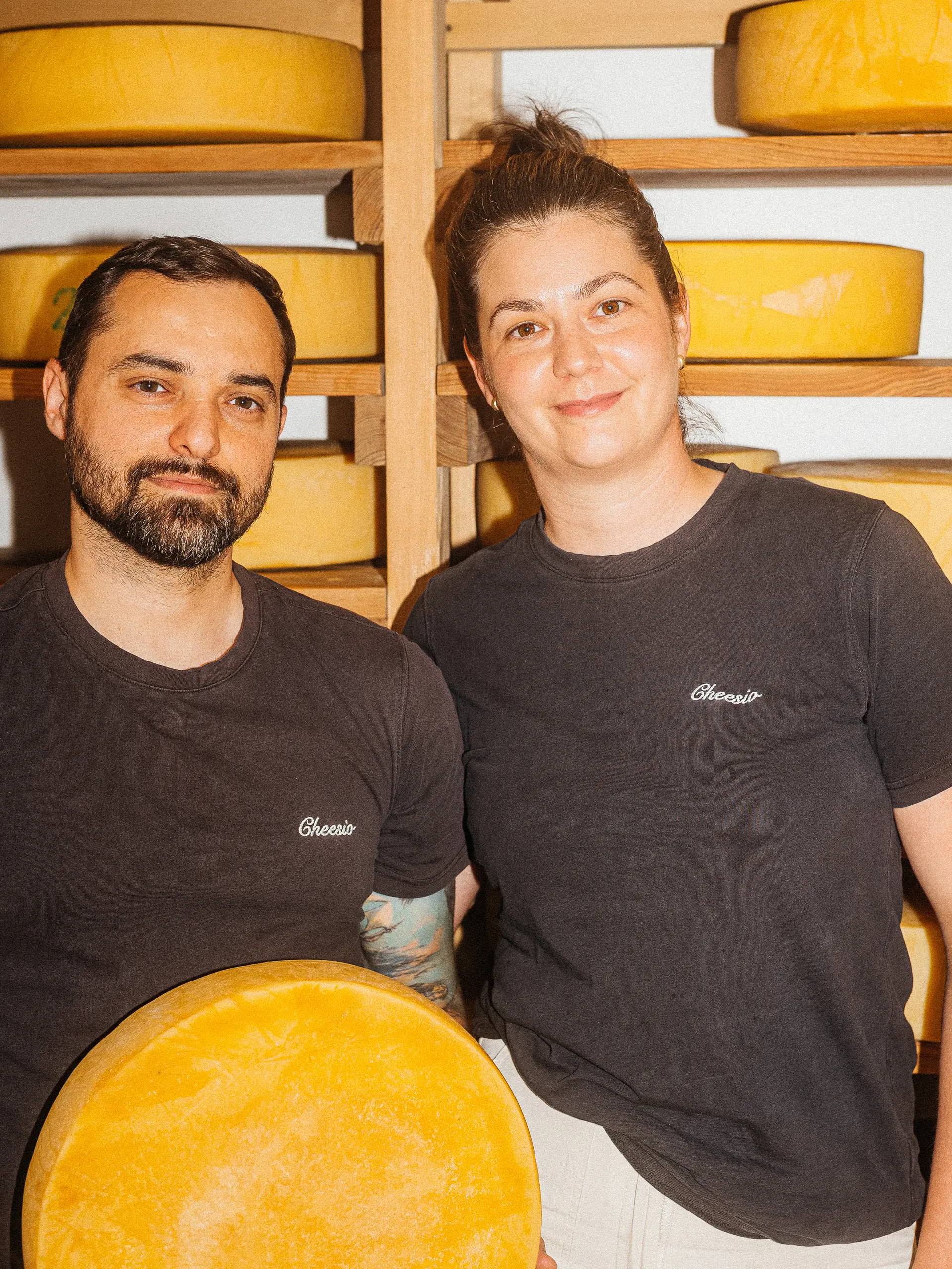Two cheesemakers stand in front of shelves of ageing cheese wheels.